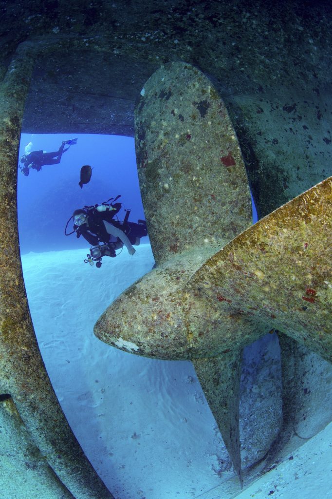 A scuba diver on a deep sea shipwreck, looking into the propeller shaft at the huge angled blades.